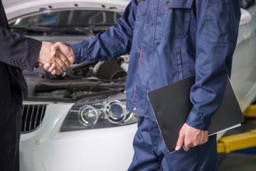Auto technician in blue coveralls shaking hands with customer in front of white BMW in repair shop.