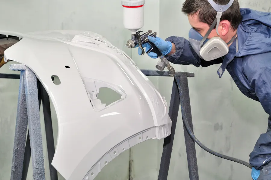 Auto body technician wearing protective gear spray painting a vehicle bumper in a professional paint booth.